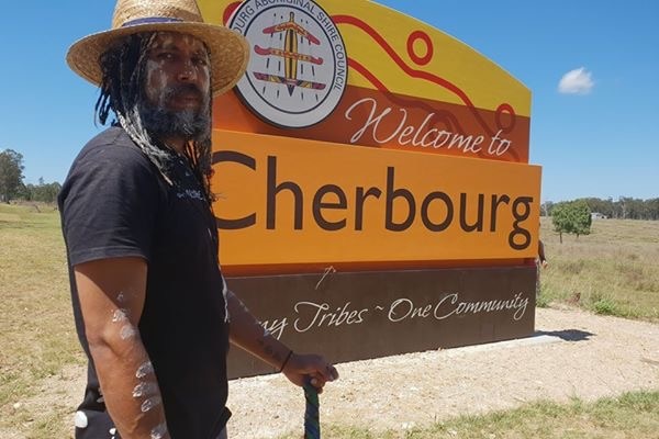 An Indigenous man wearing a straw hat stands next to a sign saying "Welcome to Cherbourg".