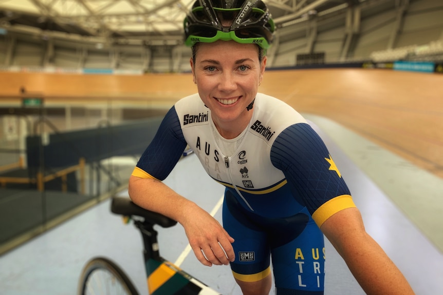 Young woman bends over her bike smiling in Olympic racing uniform at velodrome.