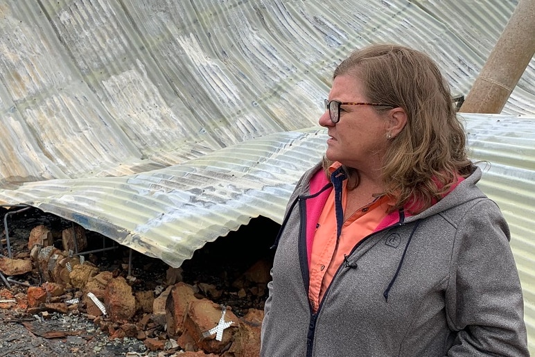 A woman stands in front of a pile of twisted corrugated iron and debris which is what remains of her home.