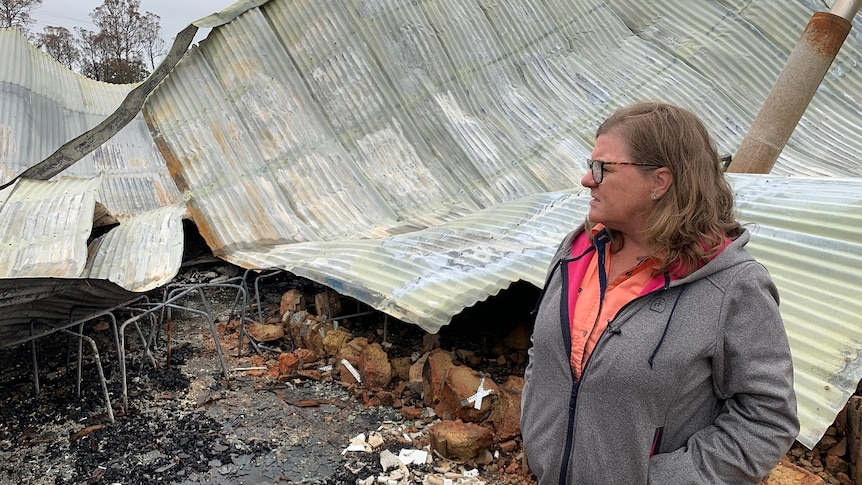 A woman stands in front of a pile of twisted corrugated iron and debris which is what remains of her home.