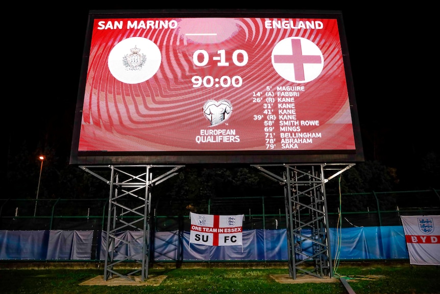 A big screen at a football ground shows the scoreline for a World Cup qualifier and the long list of England goalscorers.
