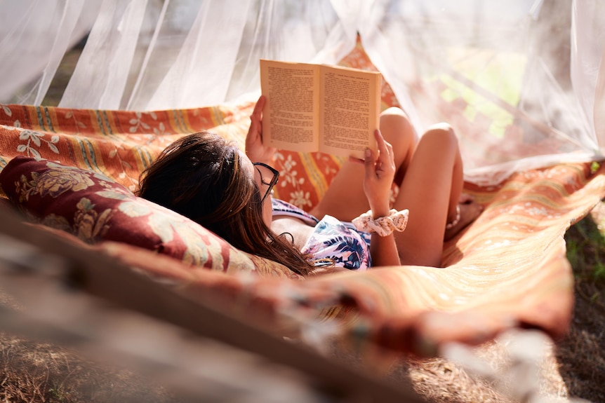 Modern woman with glasses reading a book in a hanging hammock at sunset seen from behind.