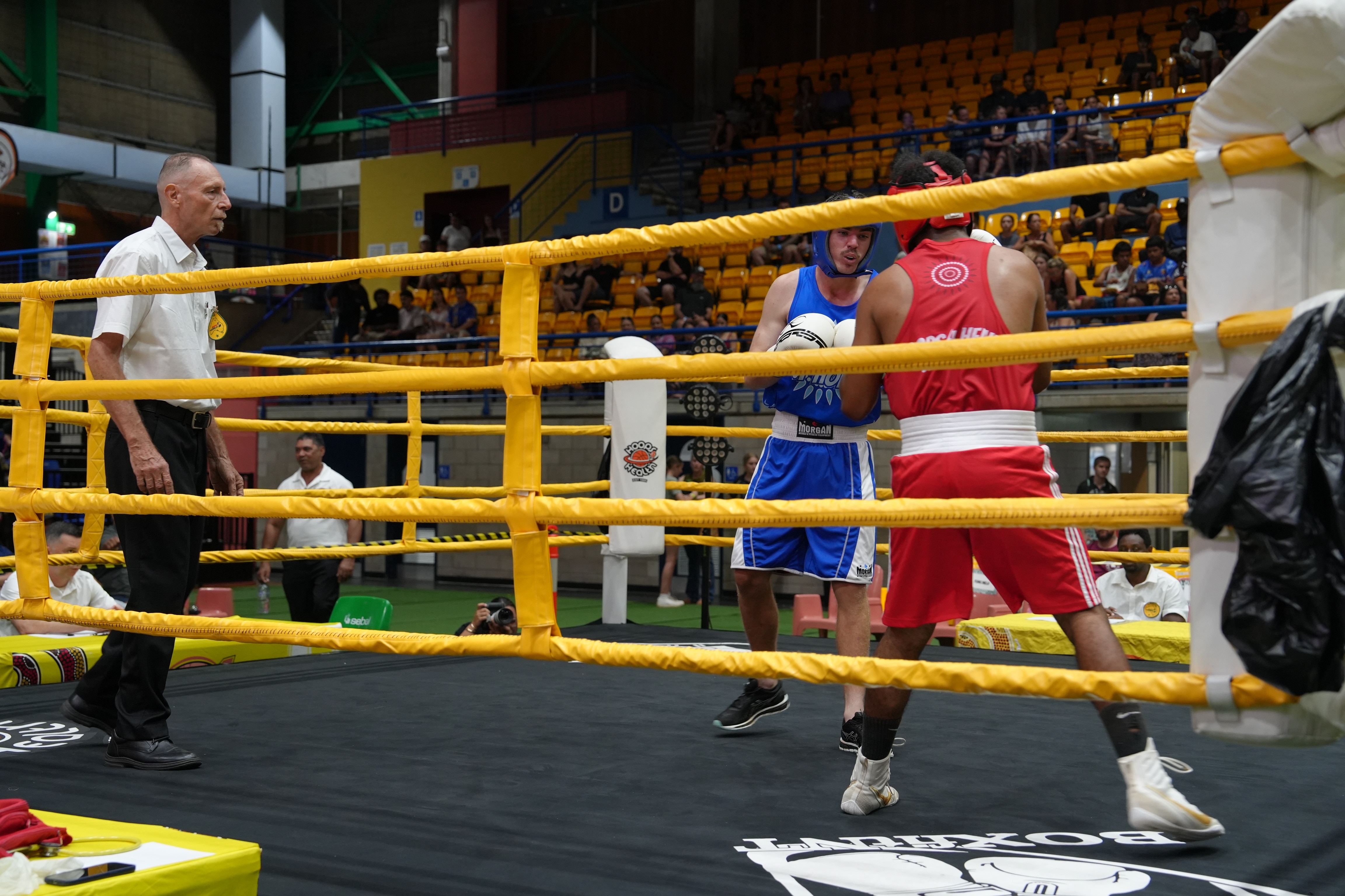 Two boxers wearing headgear compete inside a ring, one is wearing blue and the other is dressed in red.