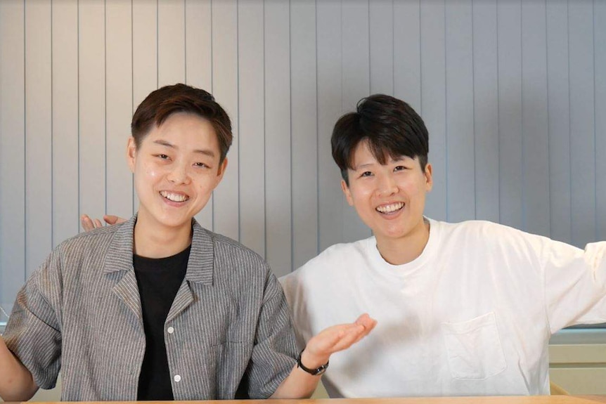 Baeck Ha-na, left, and Jung Se-young, right, smile as they look into the camera while sat behind a table. Their arms are raised.