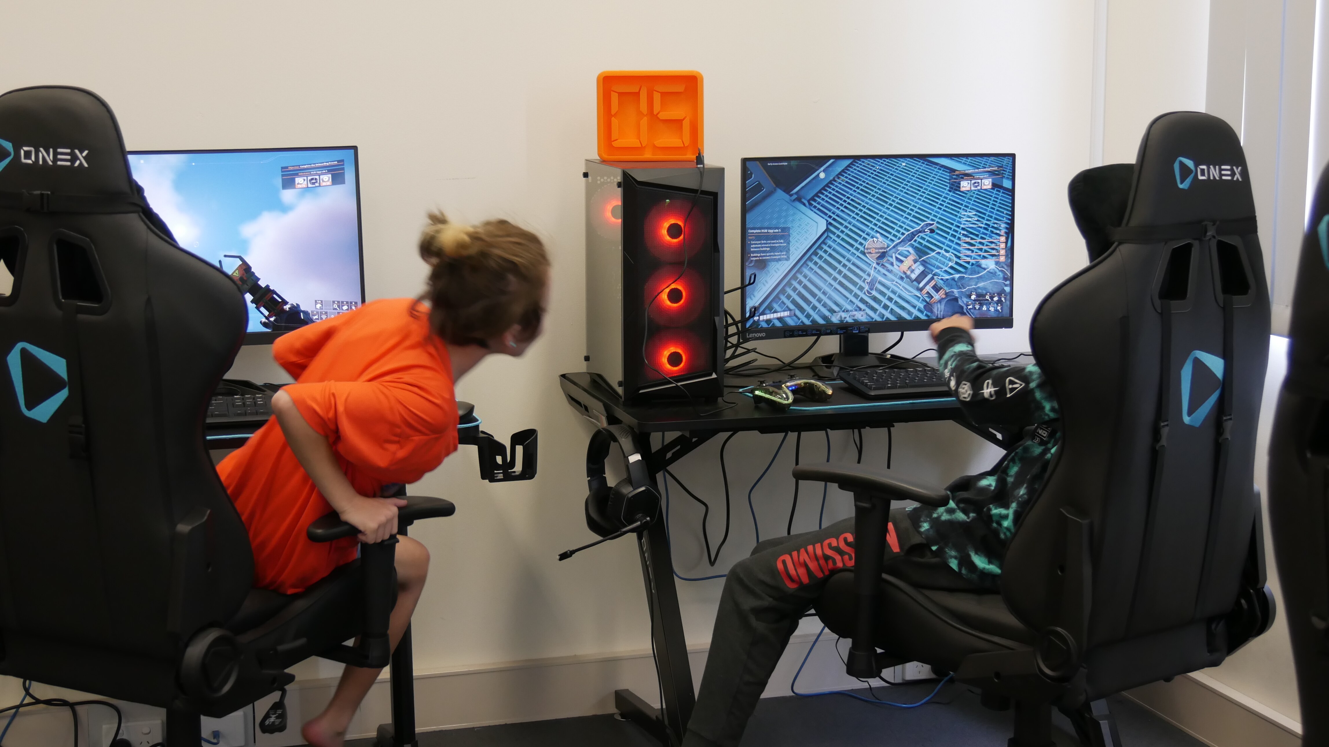 A boy in an orange tshirt leans out of his chair to look at a computer screen while a boy in a long top and pants plays a game.