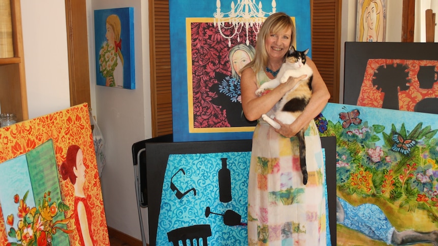 Christabel is holding a calico cats in her arms and is standing in a room filled with her colourful paintings.