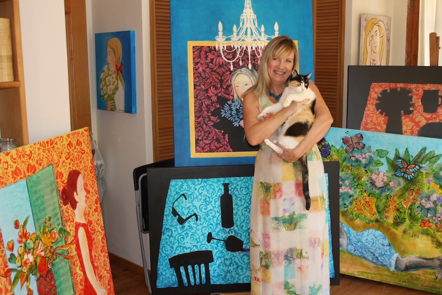 Christabel is holding a calico cats in her arms and is standing in a room filled with her colourful paintings.