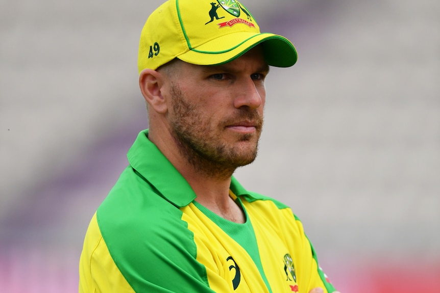 Aaron Finch stands with his arms folded, wearing yellow cricket kit