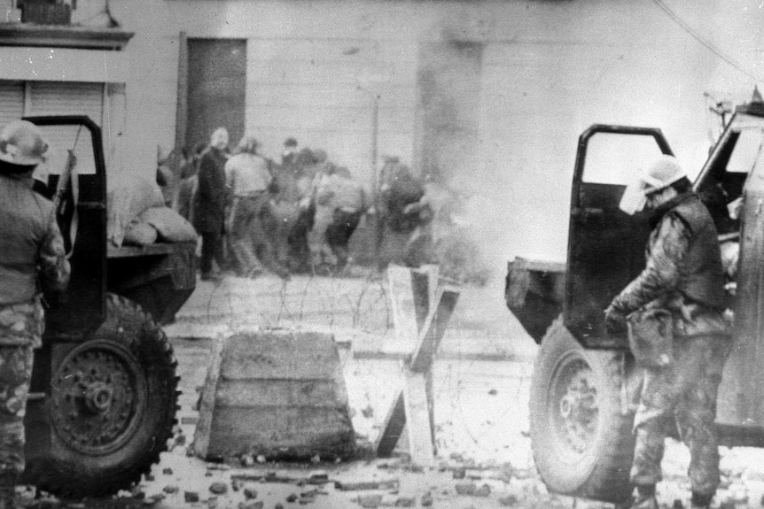 A grainy black-and-white photo from 1972 shows two soldiers in cars, barbed wire and rubble as people crouch in the background