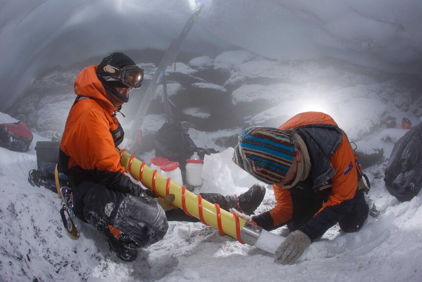 Two men in orange snow-suits, beanies, face-coverings and protective glasses, drill into ice with a cylindrical yellow drill.