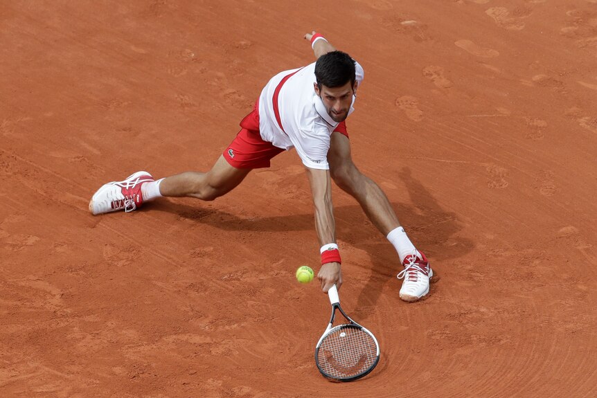 A man stands with his legs wide apart on a red clay surface with his tennis racket on the ground with a ball above