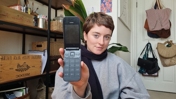 A young woman with short brown hair smiling at the camera holding up an old style flip phone.