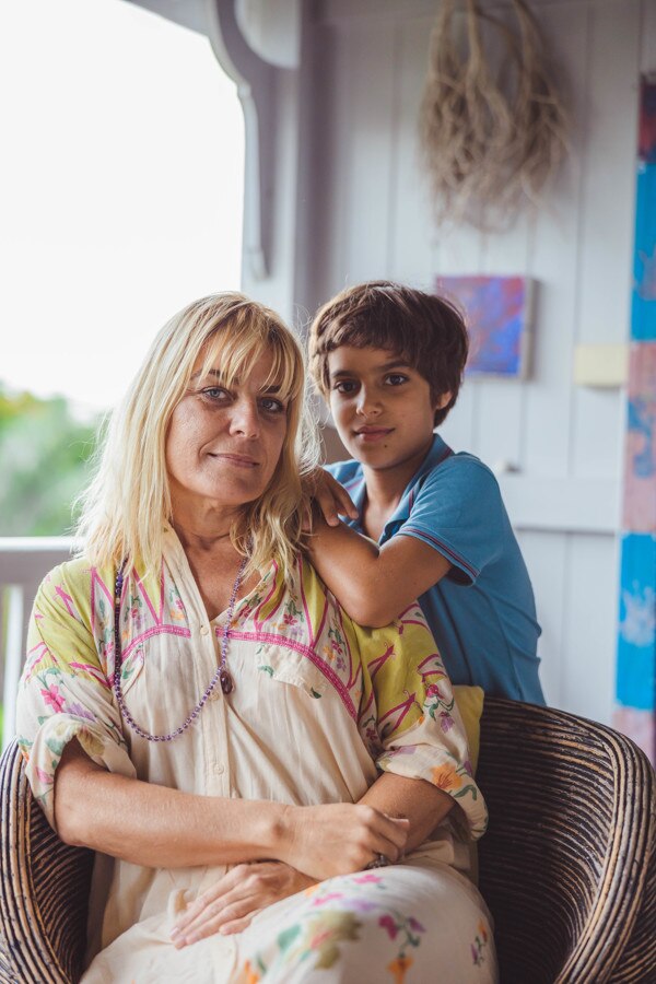 A woman is sitting on a deck at her house, looking directly at the camera, with her younger son standing next to her.