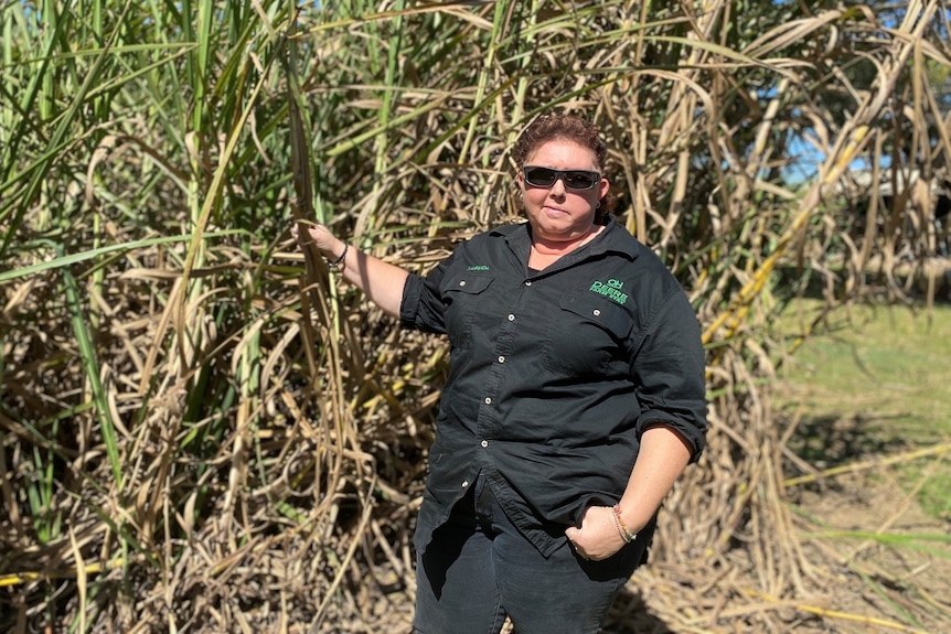 A short-haired woman wearing black shirt, pants, in front of a cane crop on her property, her hand holds onto a stalk of cane.