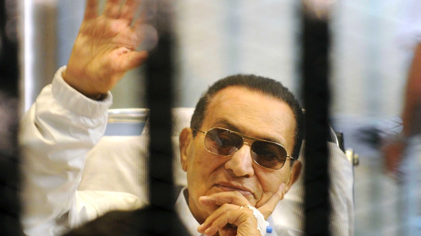 Ousted Egyptian president Hosni Mubarak waves from behind bars wearing brown aviator-style sunglasses.