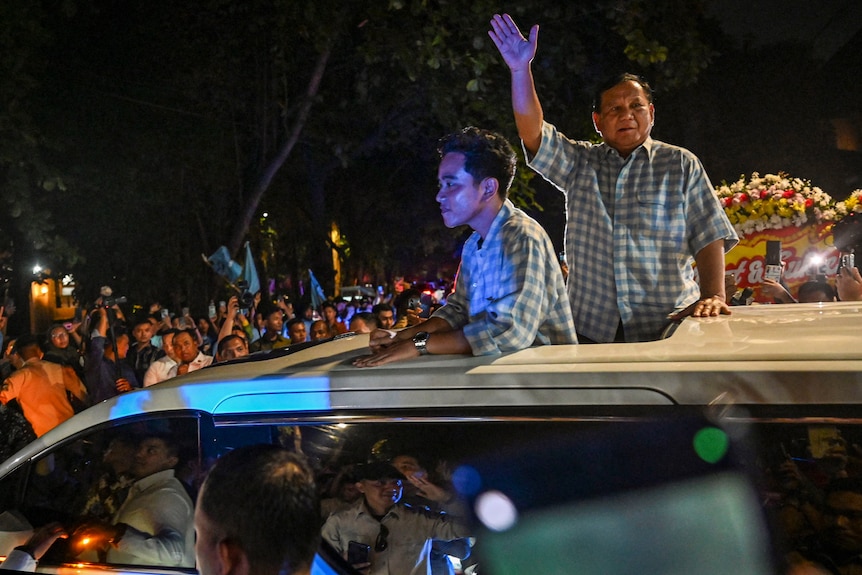 Prabowo Subianto and Gibran Rakabuming Raka stand in the sunroof of a car surrounded by supporters.