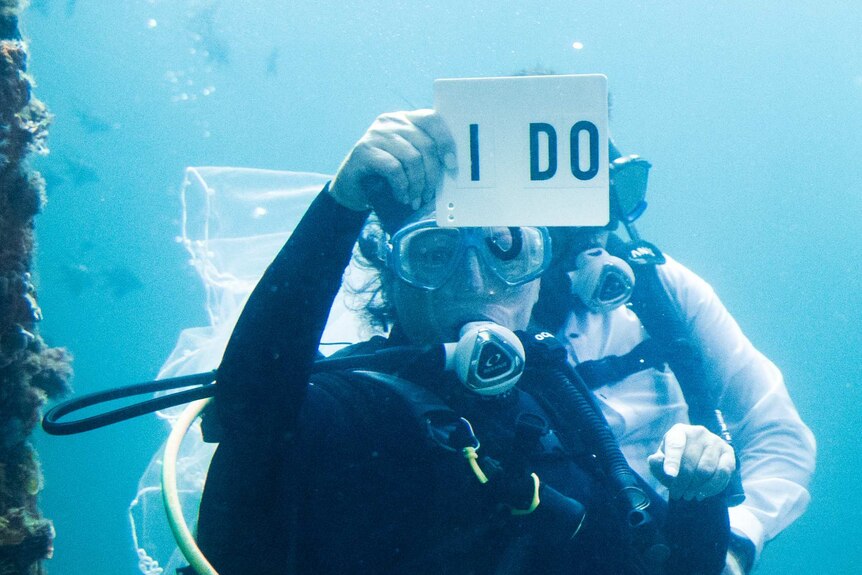 Man and woman in scuba gear underwater, woman holding 'I do' cue card