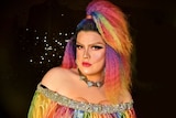 Ziggy is in drag makeup wearing a rainbow wig with a high ponytail and a rainbow off the shoulder top.
