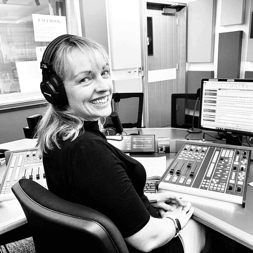 Smiling woman sitting in radio studio in front of desk of controls and microphone.
