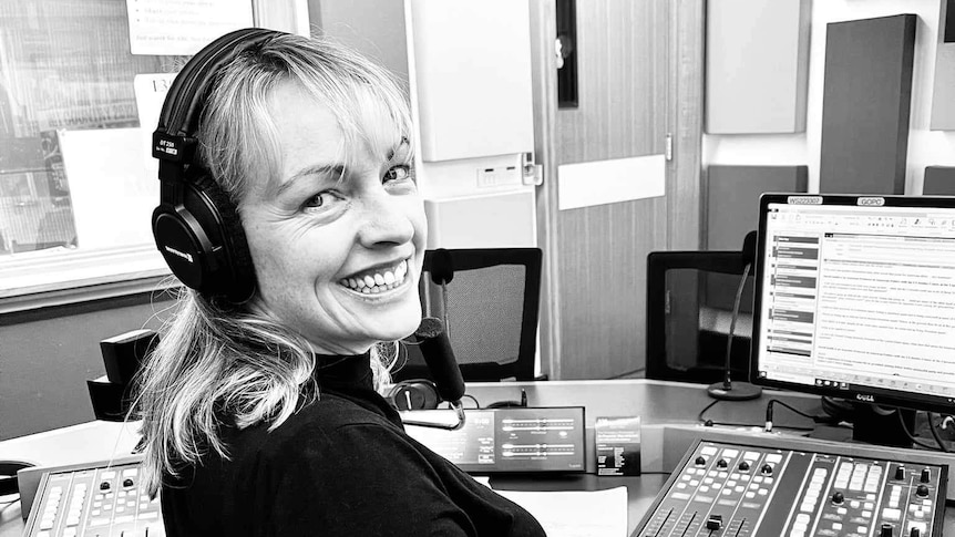 Smiling woman sitting in radio studio in front of desk of controls and microphone.