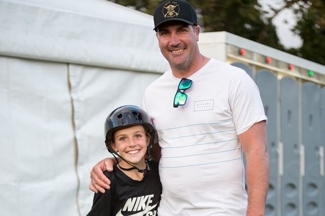 A young child to the left wearing black helmet smiling next to tall man wearing hat, white shirt and khaki pants at a skate comp