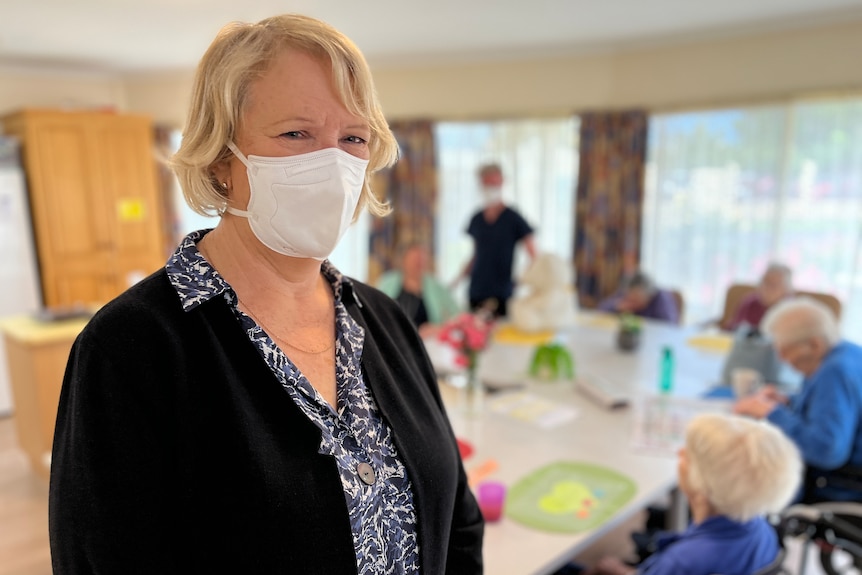Jenny Garrone wearing a face mask, with a dining table surrounded by aged care residents in the background.