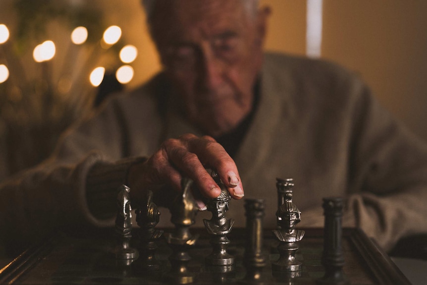 An elderly man plays chess with a set of bronzed pieces.