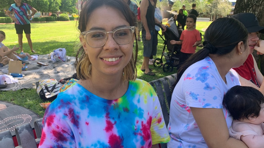 A young woman in glasses and a tie-dye shirt sitting on a park bench next to a woman holding a baby