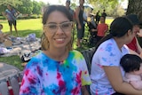 A young woman in glasses and a tie-dye shirt sitting on a park bench next to a woman holding a baby