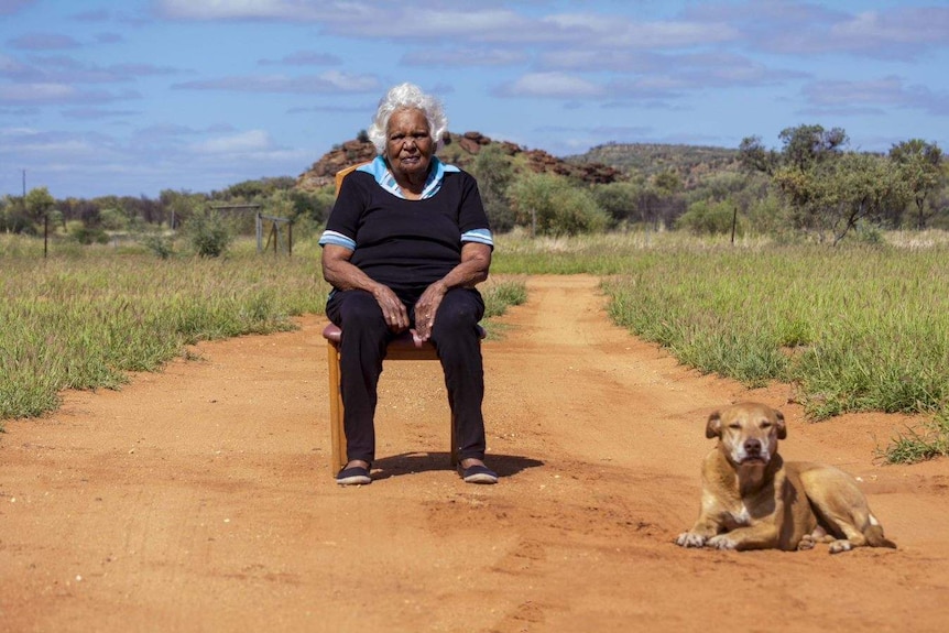 Elderly aboriginal woman sits in a chair next to her dog on a dirt track in the outback.