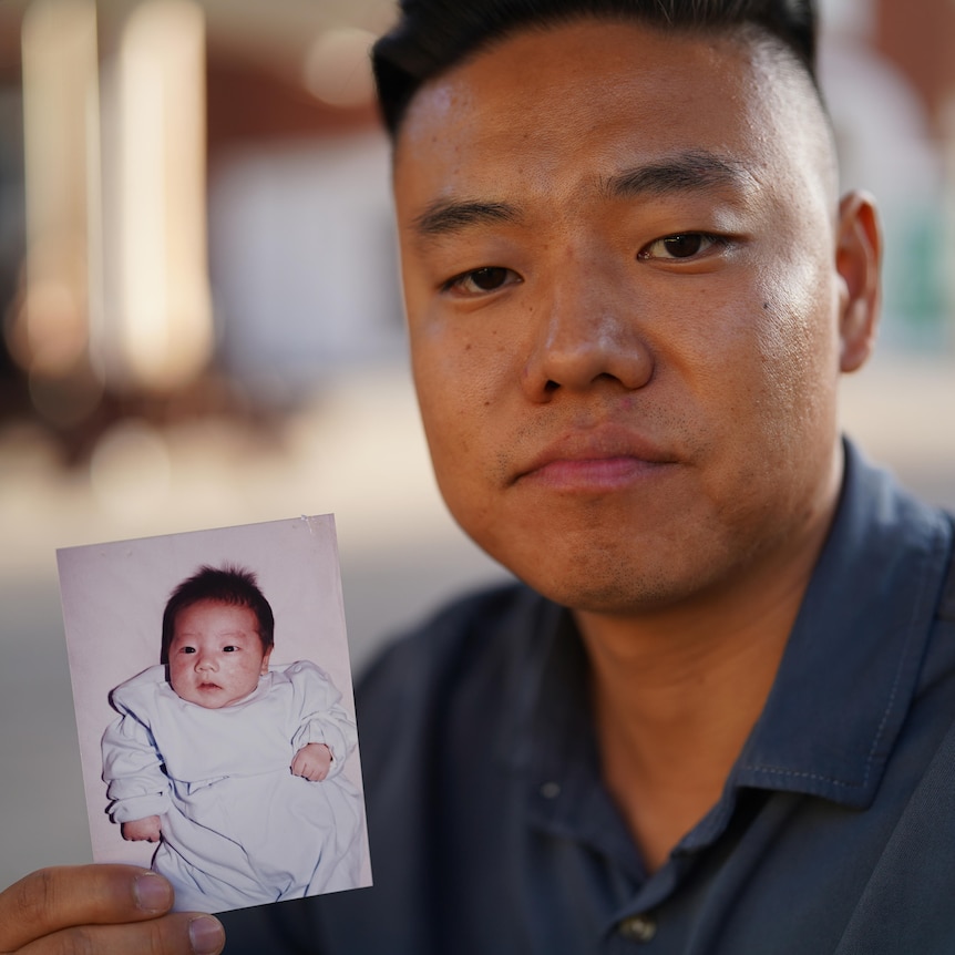 Korean man holds up a photo of himself as a baby, serious expression on his face 