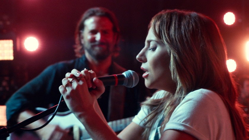 Lady Gaga holding microphone and singing into it, Bradley Cooper in background watching, red stage lighting.