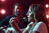 Lady Gaga holding microphone and singing into it, Bradley Cooper in background watching, red stage lighting.