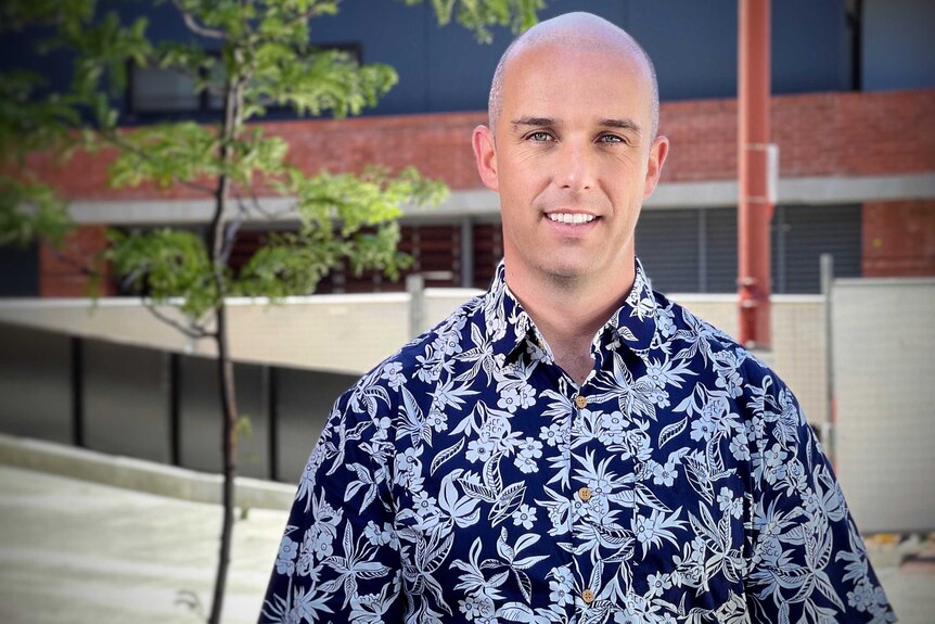 Man with shaved head and Hawaiian shirt stands in front of building and tree.