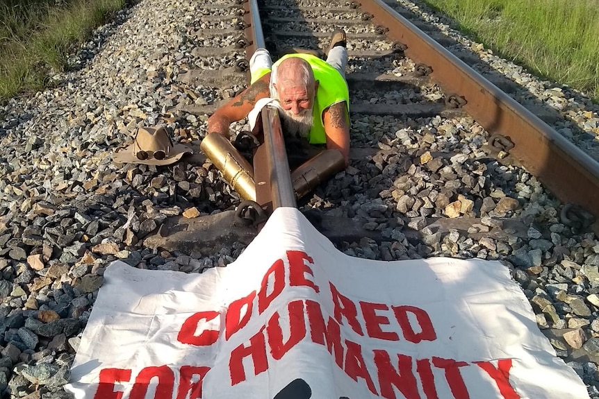 An older fellow chained to a train track with an anti-coal banner spread out in front of him.