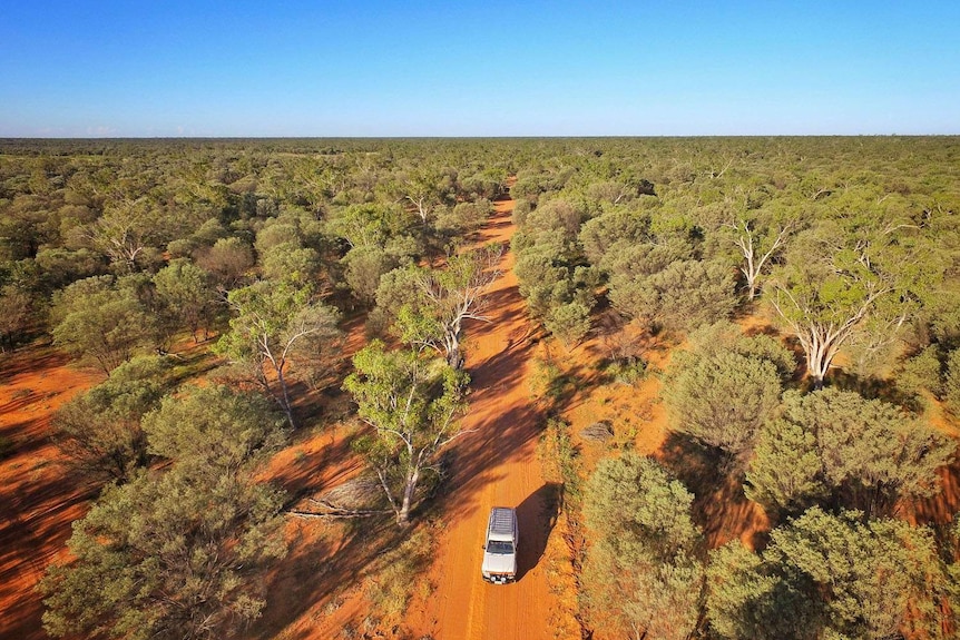A drone photo of the four-wheel-drive vehicle driving along a remote red dirt road in bushland in outback Queensland.