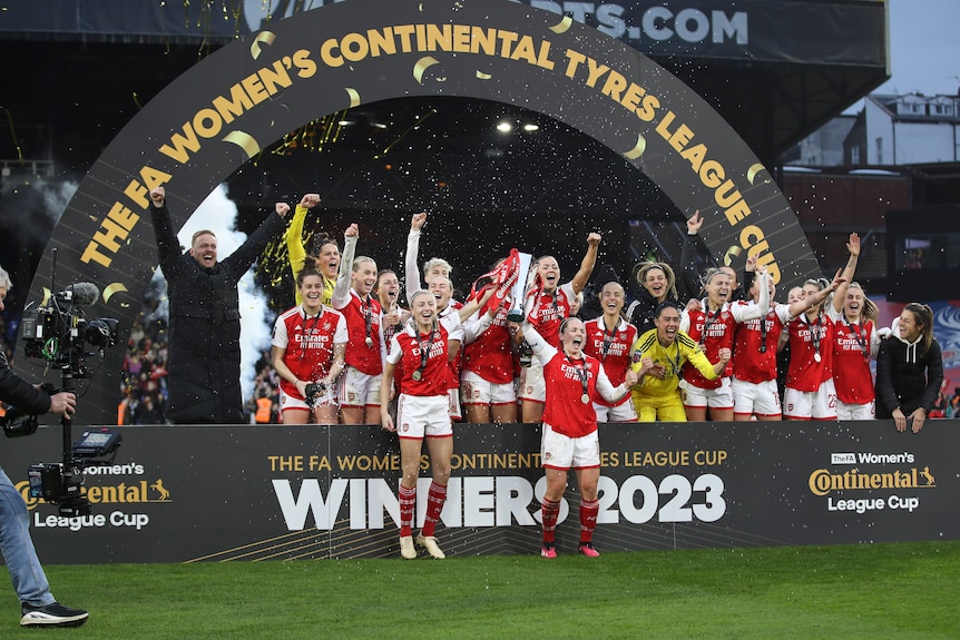 A football team in red and white holds a trophy and celebrates under a sign of a major cup competition