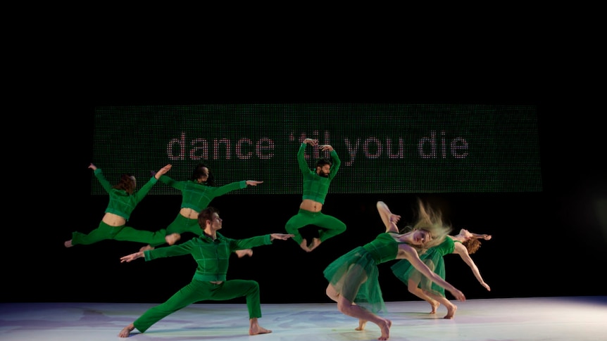 A group of dancers in green outfits dancing on a stage with the words "dance 'til you die" in the background