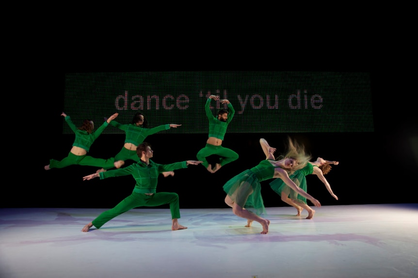 A group of dancers in green outfits dancing on a stage with the words "dance 'til you die" in the background