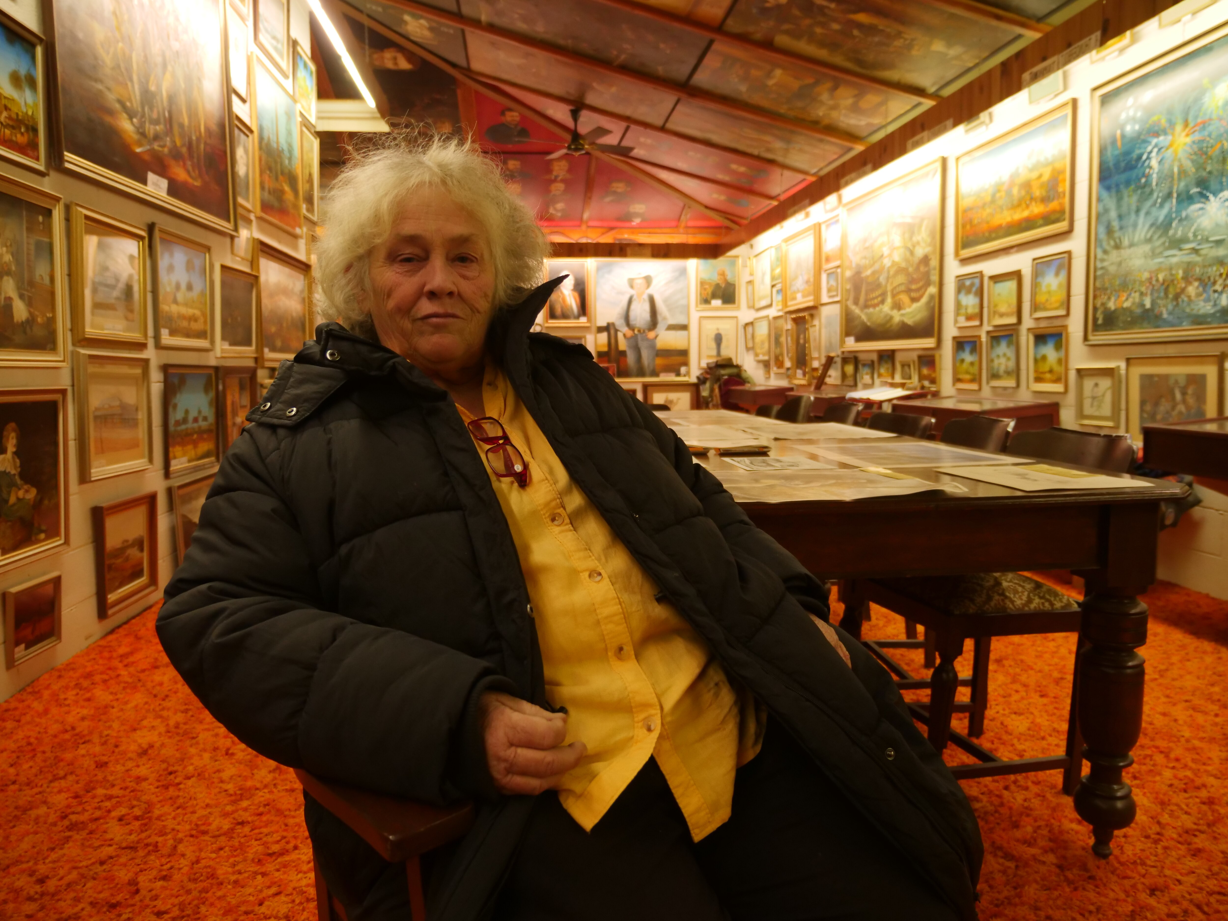 A woman wearing a big coat looks comfortable inside an art allergy with paintings covering the walls and ceiling.