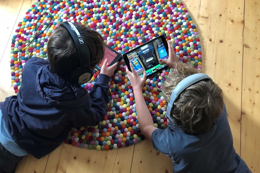 Two boys play on their iPads while wearing headphones.