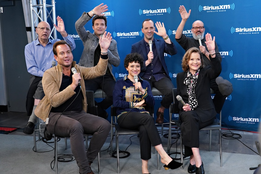 Cast sitting on chairs with hands raised, holding mics, with blue backing-board behind them.