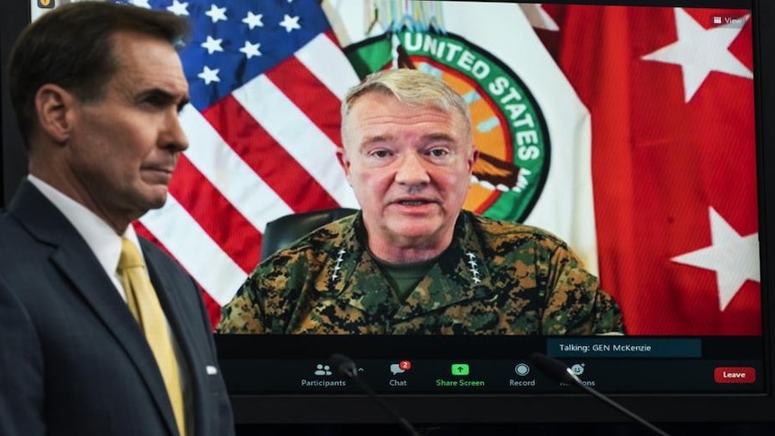 A man in the foreground wearing a suit looking off screen with pentagon official on zoom call in background 