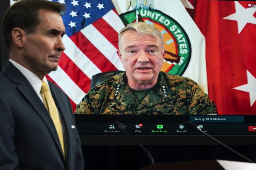 A man in the foreground wearing a suit looking off screen with pentagon official on zoom call in background 