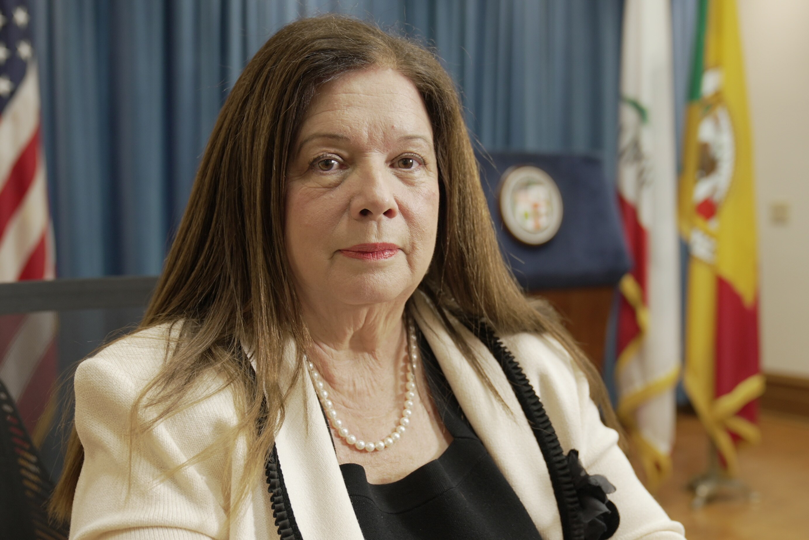 A woman wearing a black top and cream jacket with long brown hair. She also wears a pearl necklace.