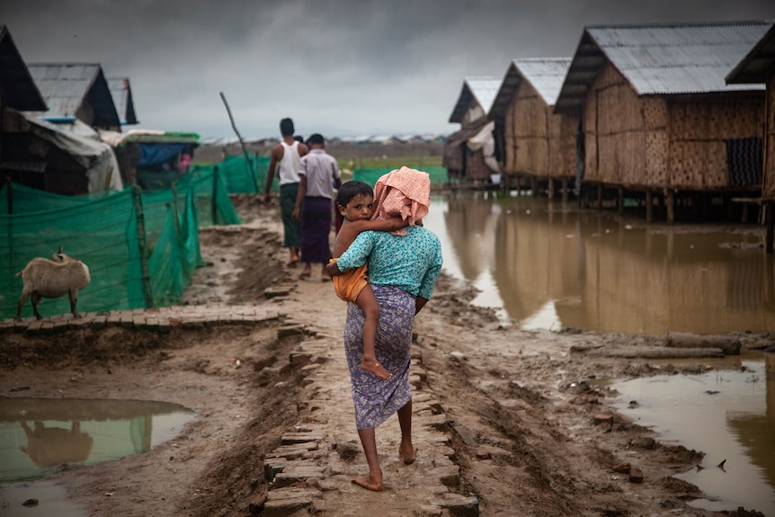 Woman walking away from camera carrying a boy looking back at the camera, surrounded by muddy water.