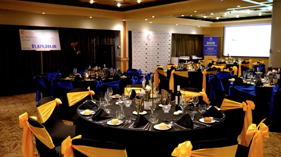 A large room with circular tables with seats decorated with yellow bows and a cheque in the background.