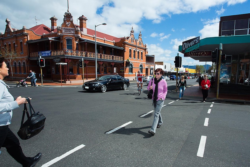 Pedestrians and cars navigate an intersection in a town centre with grand brick hotel on the corner.