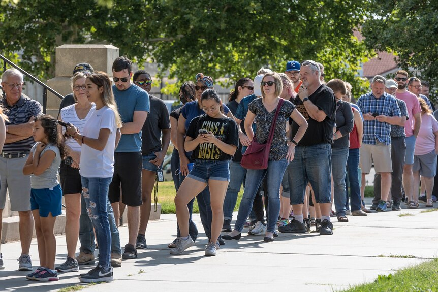 A long line of people standing on a footpath - most dressed in t-shirts and shorts and caps