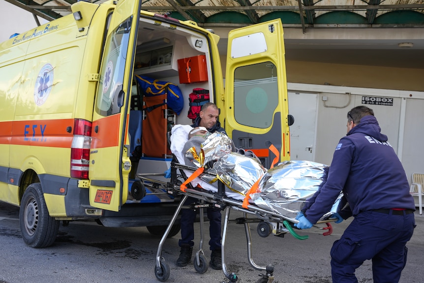 An injured man wrapped in aluminium foil on a stretcher being carried by two paramedics into a yellow ambulance